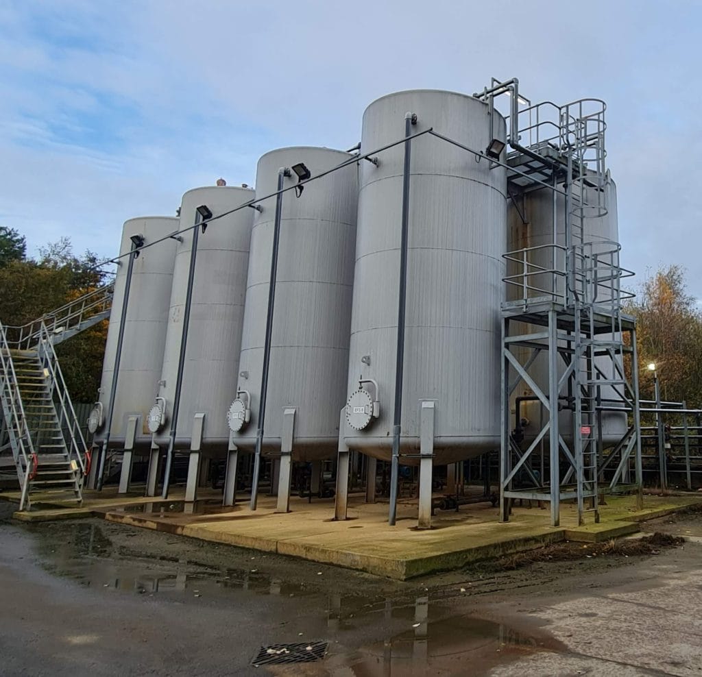 Four large cylindrical industrial storage tanks are positioned side by side on a concrete platform, with metal stairs and railings providing access. Regular non-destructive testing (NDT) ensures the safety and integrity of these vital storage units.
