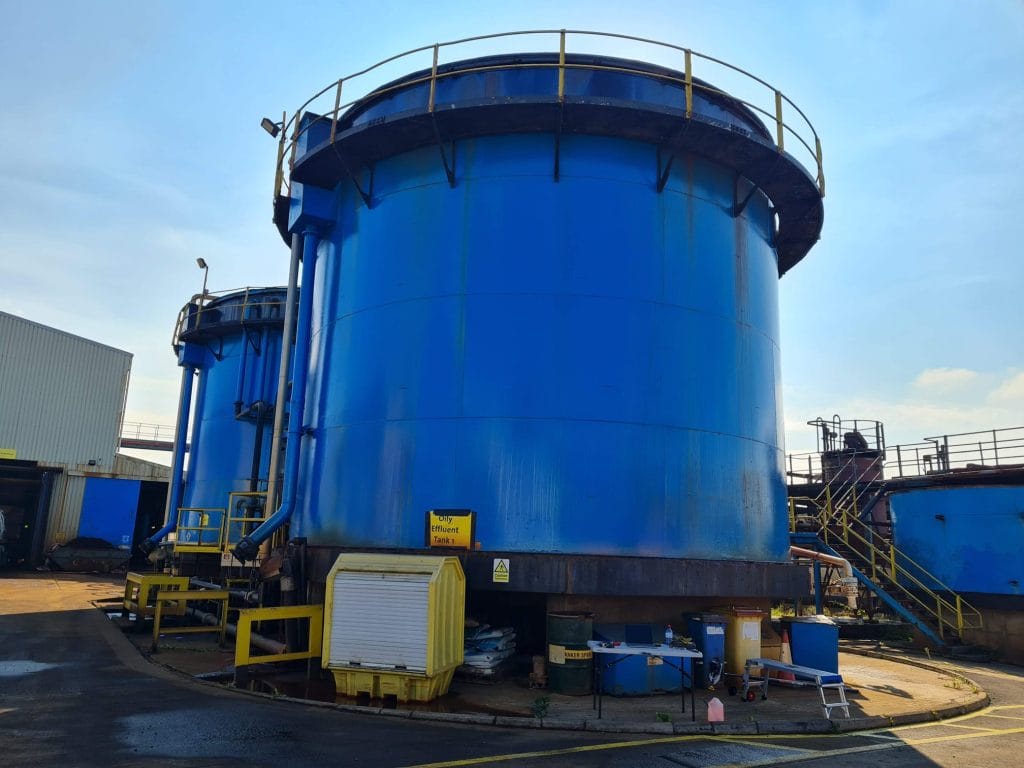 Large cylindrical blue industrial storage tanks with yellow railings and steps, located outdoors at an industrial facility under a clear sky, where routine non-destructive testing (NDT) ensures safety and structural integrity.