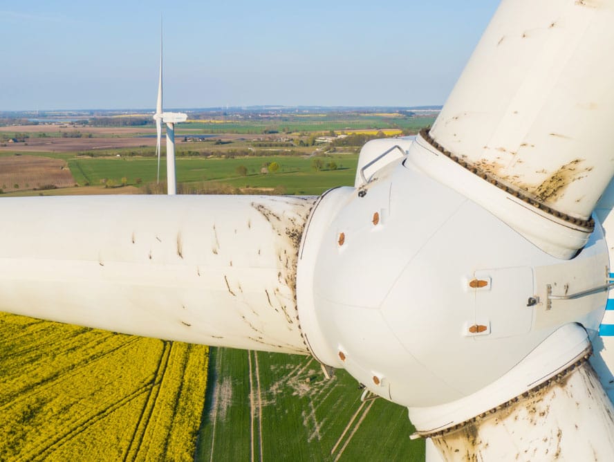 Close-up view of a wind turbine rotor with blades, set against a rural landscape with fields and another wind turbine in the background, highlighting areas where non-destructive testing (NDT) ensures structural integrity.
