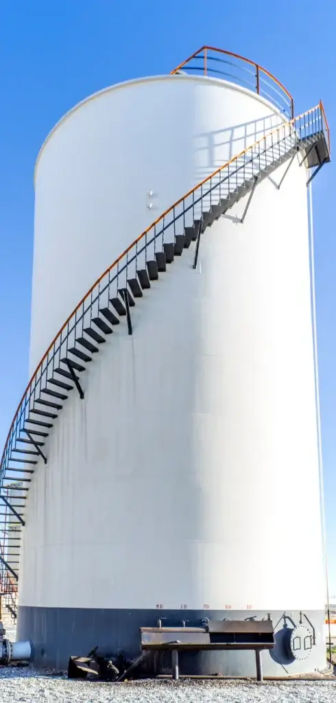 Large white industrial storage tank with a black spiral staircase wrapping around its exterior, set against a clear blue sky—a prime candidate for regular non-destructive testing (NDT) to ensure safety and structural integrity.