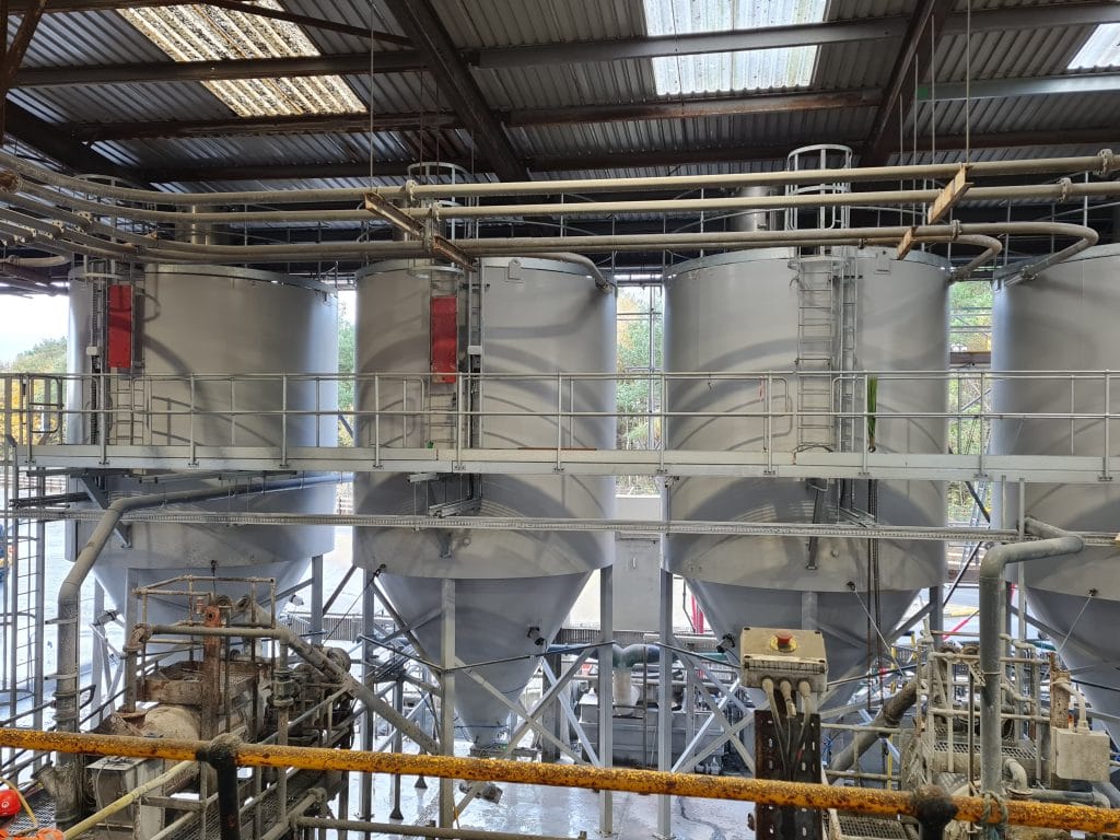Four large industrial storage silos with metal walkways and pipes inside a warehouse facility, where machinery and equipment support non-destructive testing (NDT) operations.